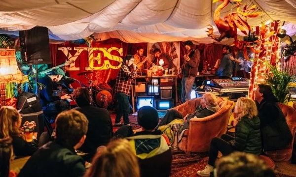 A lively indoor music performance with musicians, an audience seated on plush furniture, colorful decorations, and atmospheric lighting creating a cozy ambiance.