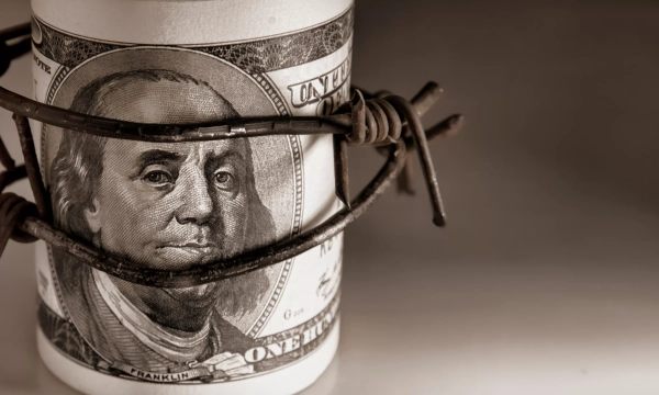 A rolled-up hundred-dollar bill featuring Benjamin Franklin is tightly bound by barbed wire, symbolizing restraint or entrapment related to money.