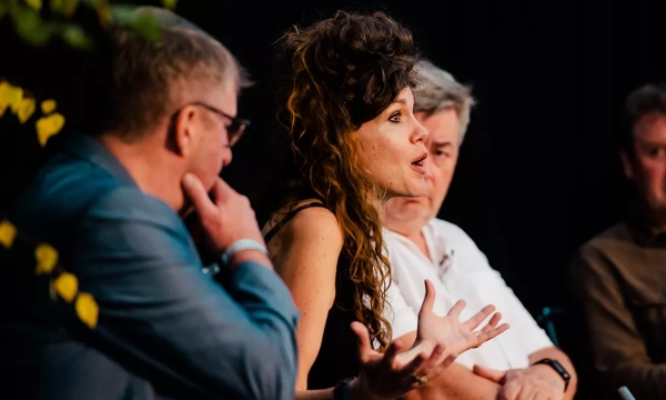 A woman passionately speaking during a panel discussion, flanked by three men who are attentively listening. The setting features soft lighting and foliage in the foreground.