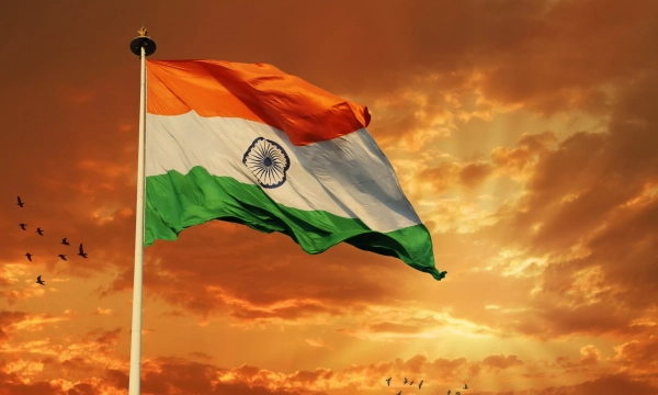 The Indian national flag waving against a dramatic sunset sky, with birds flying in the background.