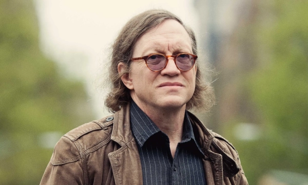 A middle-aged man with long, wavy hair and round sunglasses stands outdoors, wearing a brown jacket and a black shirt, with greenery in the background.