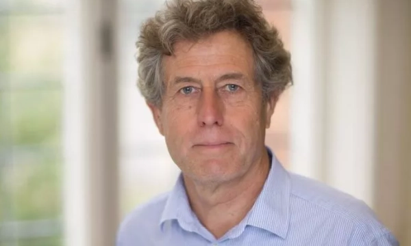 A middle-aged man with curly hair, wearing a light blue shirt, stands in a bright room with windows in the background.