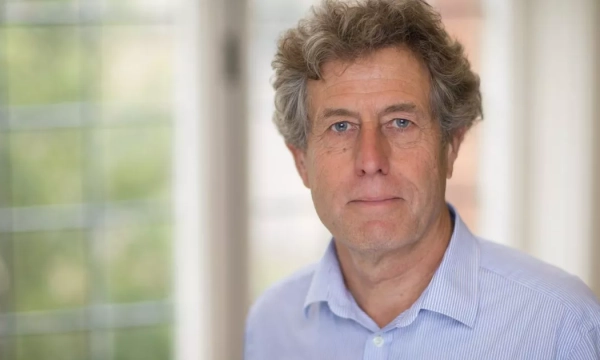 A middle-aged man with curly hair, wearing a light blue shirt, standing indoors with natural light coming through a window in the background.
