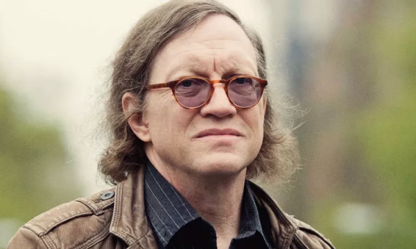 A middle-aged man with long, wavy hair and round glasses, wearing a brown leather jacket, stands outdoors with a serious expression.