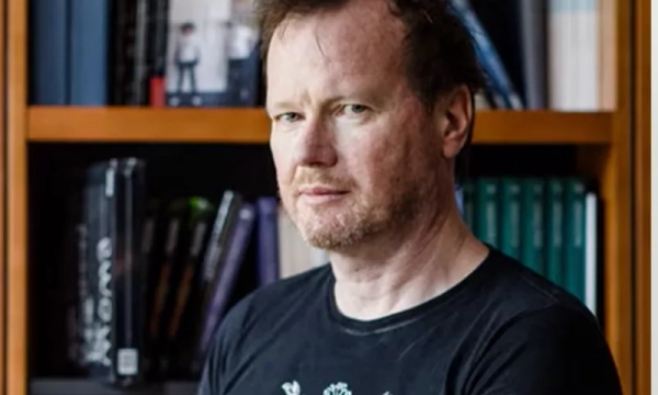 A middle-aged man with tousled hair stands in front of a bookshelf filled with books, wearing a black t-shirt.