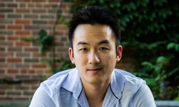 A young man with short black hair, wearing a light blue shirt, is seated outdoors in front of a brick wall and green foliage.