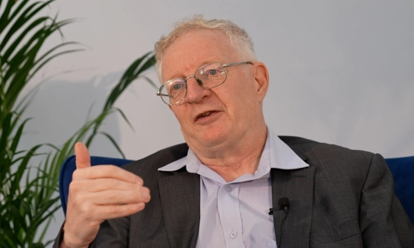 A middle-aged man with glasses and a suit gestures while speaking in an interview setting, surrounded by plants and a blue couch.