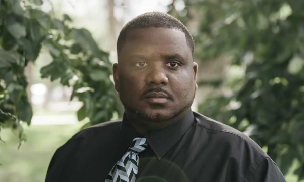 A man in a black shirt and patterned tie stands outdoors, surrounded by green foliage, with a serious expression on his face.
