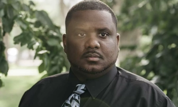 A man in a black shirt and patterned tie stands outdoors among greenery, looking directly at the camera with a serious expression.