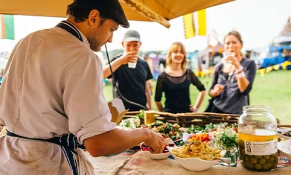 A chef prepares a dish at a food stall, while three customers watch and enjoy drinks in a vibrant outdoor market setting.