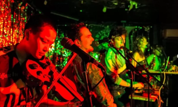 A lively band performing on stage, featuring musicians with a violin, keyboard, and vocals, illuminated by colorful stage lights and a sparkling backdrop.