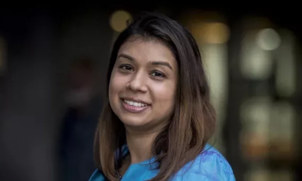 A smiling woman with brown hair and a blue shirt, standing against a blurred background, exuding a friendly demeanor.