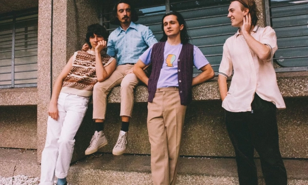 A group of four young adults, dressed in casual attire, casually posing together outside a textured concrete building. They're smiling and interacting with each other.