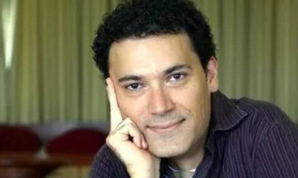 A man with curly hair rests his chin on his hand, smiling slightly as he poses for the camera. Background features soft, muted tones.