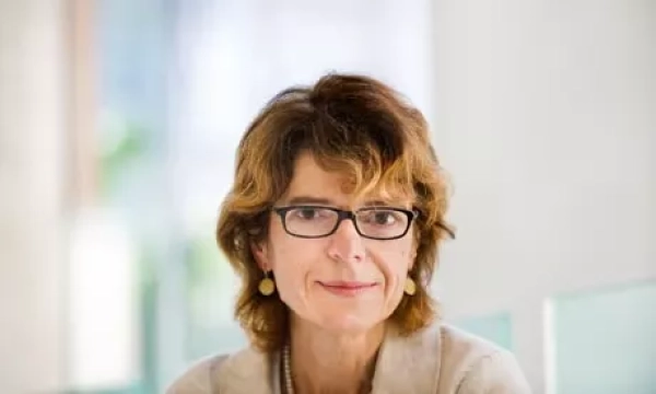 A woman with curly brown hair and glasses, wearing a light-colored sweater, sits in a well-lit indoor space, looking directly at the camera.