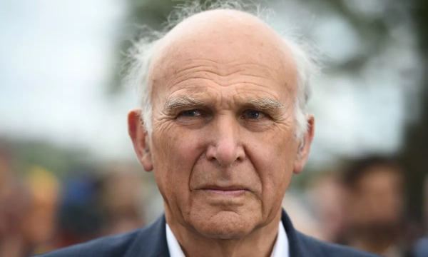A senior man with gray hair and a serious expression stands outdoors, blurred crowd in the background.