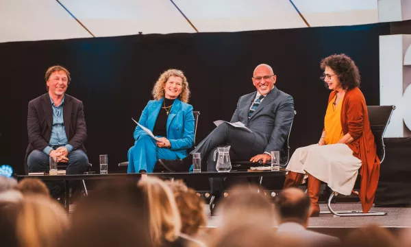 A panel discussion featuring four speakers sitting at a table, engaged and smiling, under a large tent with an enthusiastic audience in the foreground.