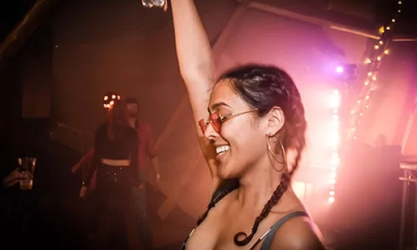 A woman with braided hair dances joyfully in a vibrant, dimly lit party scene, holding a water bottle above her head.