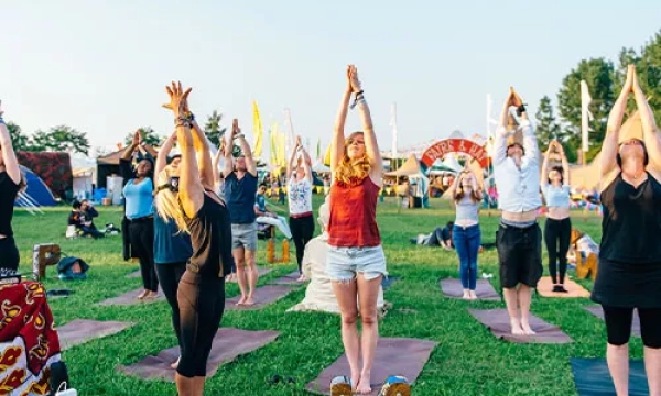A group of people practicing yoga outdoors on a grassy field, raising their arms with a colorful festival backdrop in the distance.