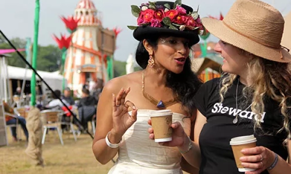 Two women engage in conversation at an outdoor festival, one wearing a floral hat and the other in a straw hat with 