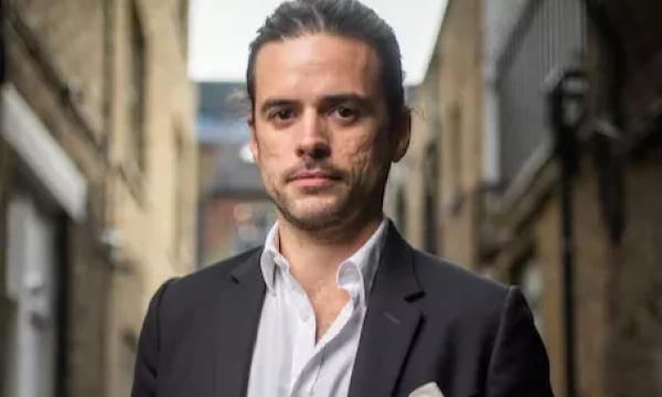 A man in a black jacket and white shirt stands in a narrow alleyway, exuding a confident demeanor.