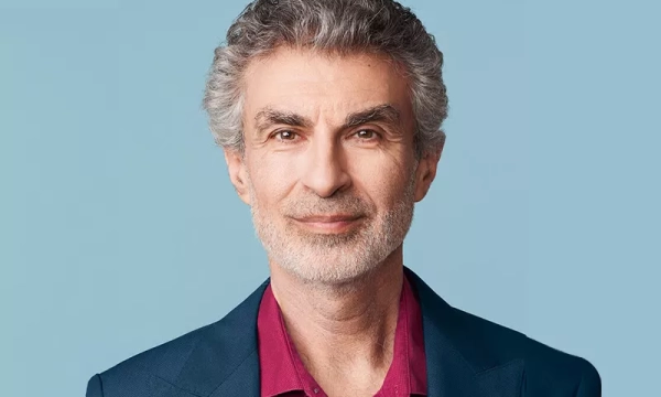 A man with gray hair and a beard, wearing a dark blazer over a red shirt, smiles against a light blue background.