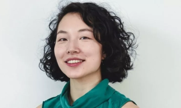 A smiling woman with curly hair wearing a teal top against a plain background. She has a confident expression and subtle makeup.