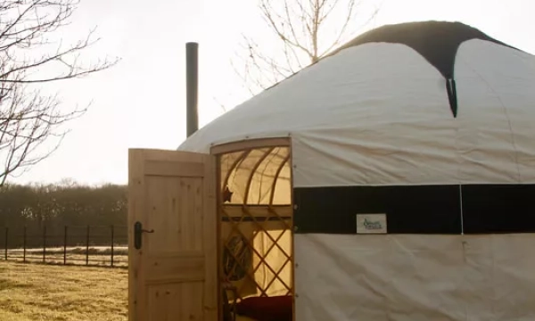 A wooden door leads into a white yurt with a black band, set in a grassy area with trees in the background under a clear sky.