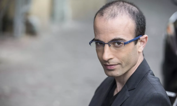 A man in a black suit and glasses gazes intently at the camera, with a blurred street background.