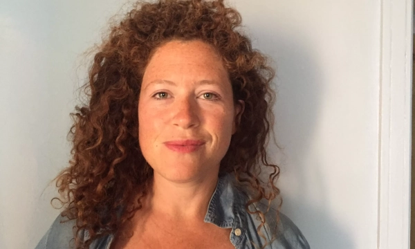 A woman with curly red hair smiles at the camera, wearing a denim shirt against a plain background.