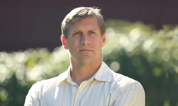 A serious-looking man with short gray hair, wearing a light striped shirt, stands outdoors with greenery in the background.
