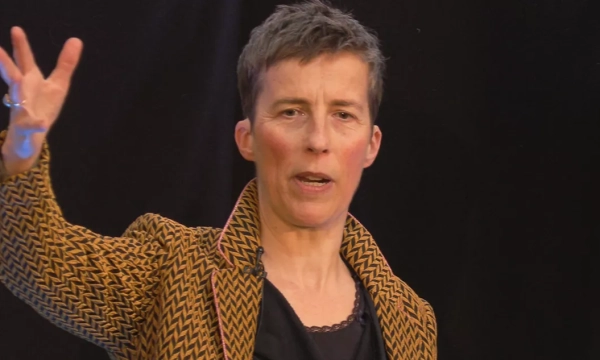 A woman with short hair and a patterned jacket gestures while speaking, conveying a sense of engagement and enthusiasm.