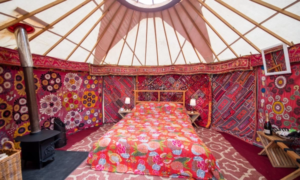 A colorful yurt interior featuring a floral-patterned bedspread, wooden bed frame, two lamps, and decorative walls, creating a cozy and vibrant atmosphere.