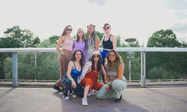 A group of seven young women poses together outdoors on a bridge, smiling and wearing summer outfits, surrounded by greenery and a partially cloudy sky.