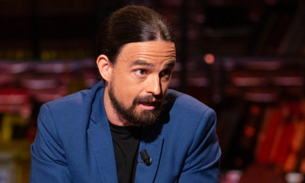 A man with long hair and a beard speaking intently while wearing a blue blazer, set against a dimly lit backdrop.