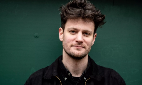 A young man with tousled dark hair and a slight smile stands against a green background, wearing a black jacket over a dark shirt.
