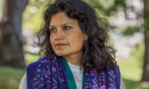 A woman with long, curly hair is outdoors, wearing a white top and a colorful scarf, looking thoughtfully into the distance amidst a blurred green background.