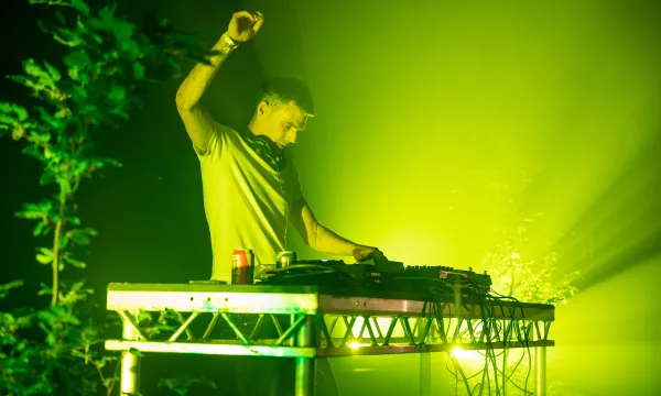 A DJ performs on stage, illuminated by green lighting, with a mixing console and sound equipment, creating an energetic atmosphere.