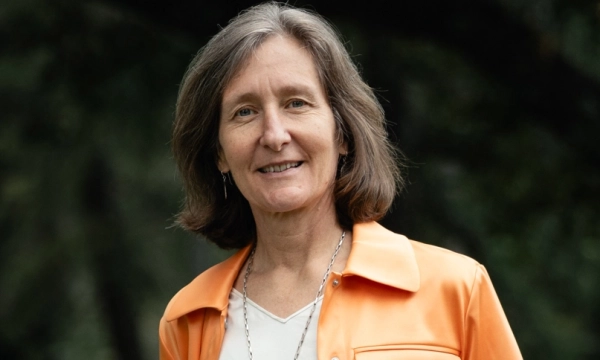 A woman with shoulder-length brown hair smiles while wearing an orange jacket over a white shirt, standing outdoors with blurred greenery in the background.