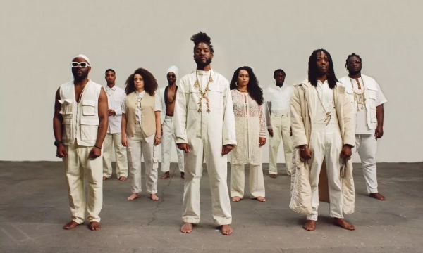 A diverse group of ten people stands barefoot in a minimalist studio, dressed in various shades of white and beige, with a focus on unity and expression.