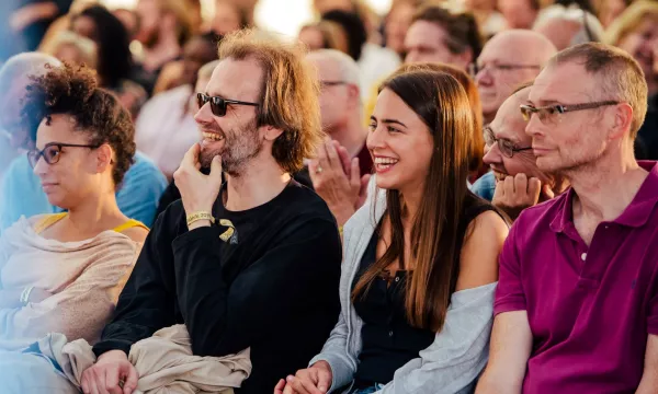 A diverse audience enjoying a performance, with a focus on a man with glasses and a woman smiling, seated among others.
