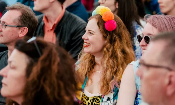 A lively audience at an event, focusing on a smiling woman with curly hair wearing a colorful pom-pom headband, surrounded by diverse attendees.