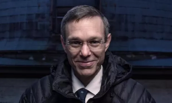 A man in a black puffer jacket and glasses smiles against a dark, industrial background.