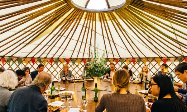 A group of people gathered around a large circular table inside a yurt, sharing a meal, with a decorative centerpiece and warm lighting.