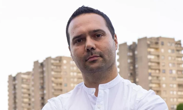 A serious-looking man in a white shirt stands with crossed arms in front of tall apartment buildings.