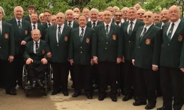 A group of men in green blazers and ties, standing together in an outdoor setting, with one individual in a wheelchair at the front.