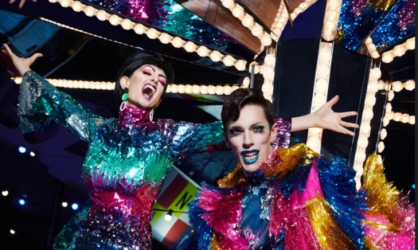 Two performers in vibrant, sequined costumes with bold makeup, posing joyfully under bright lights and colorful decorations, emphasizing a lively and festive atmosphere.