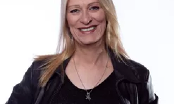 A smiling person with long blonde hair, wearing a black leather jacket and a dark top, stands against a white background.