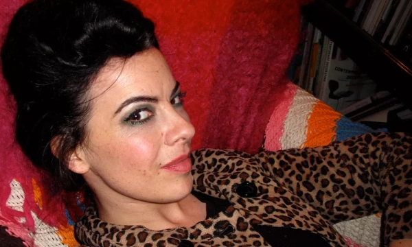 A woman with dark hair styled in an updo, wearing a leopard print coat, reclines comfortably against a colorful knitted blanket.