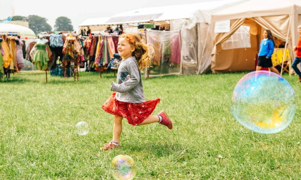 A joyful young girl with curly red hair runs through a grassy field, surrounded by colorful bubbles and outdoor vendor tents.